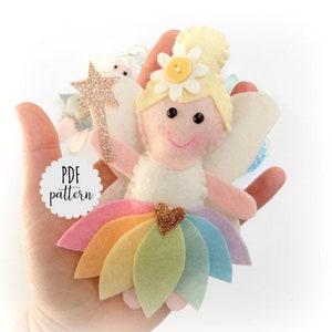 May include: A felt rainbow fairy doll with a white dress, yellow hair, and a gold glitter wand. The doll has a white flower with a yellow button in the center on her head. The doll is holding the wand in her right hand. The doll is wearing a rainbow skirt with a gold glitter heart in the center. The doll is sitting on a hand. The text "PDF pattern" is on a white circle with a scalloped edge.
