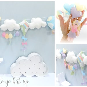 May include: A pastel-coloured felt garland featuring clouds, hot air balloons, and a small fairy doll. The garland is strung on a white string and is hanging on a light blue wall. The text "There's nowhere to go but up" is written below the garland.