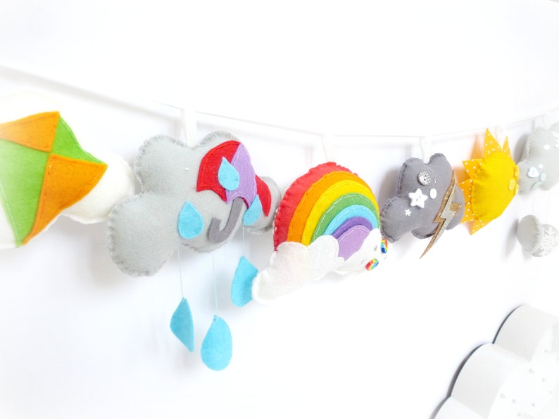 K&ouml;nnte beinhalten: Eine Filz-Girlande mit einem Regenbogen, Wolken, einer Sonne, einem Drachen und einer Gewitterwolke mit Blitz. Die Girlande ist an einer wei&szlig;en Schnur befestigt und eignet sich perfekt f&uuml;r ein Kinderzimmer oder ein Babyzimmer.
