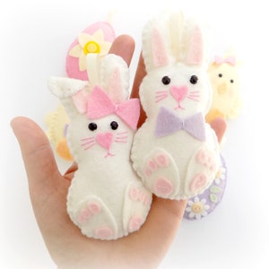 May include: Two white felt bunny ornaments with pink and purple bows. The bunnies have black eyes and pink noses. The ornaments are hanging from a hand.