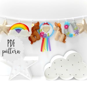 May include: A felt banner with a rainbow, two horses, a horseshoe, two stars, and a heart with a glitter finish. The banner is strung on a white ribbon. The text "PDF pattern" is on a white circle.
