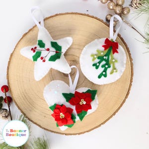May include: Three felt Christmas decorations, a star, a circle, and a heart, each decorated with green holly, red berries, and white beads. The decorations are white with red and green accents.