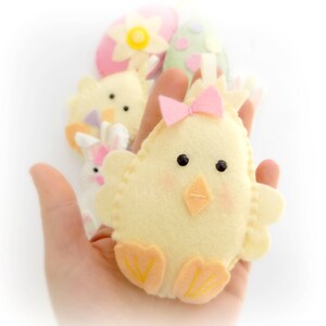 May include: A yellow felt chick with a pink bow, orange feet, and black eyes. The chick is held in a hand.