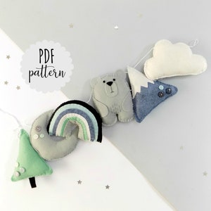 May include: PDF pattern for a felt garland with a rainbow, a mountain, a cloud, a bear, and a tree. The garland is made of felt and has a white background.