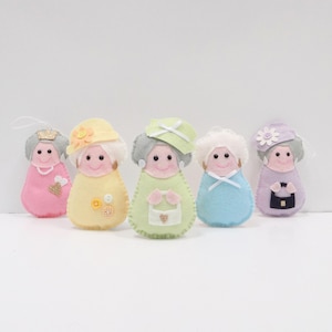 May include: Five felt ornaments shaped like people, each with a different color and design. The ornaments are all wearing hats and have smiling faces.