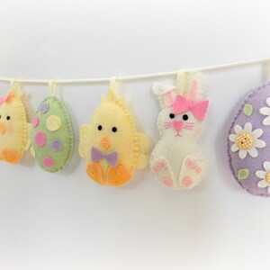 May include: A felt Easter garland featuring a bunny, chicks, and eggs. The garland is decorated in pastel colours and has a pink bow on the bunny.