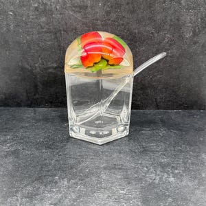 May include: A clear, hexagonal glass jar with a lid featuring a red pepper design. A clear spoon rests inside the jar. The jar is on a dark grey surface.