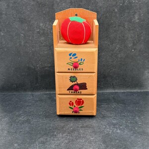 May include: Wooden sewing organiser with three drawers labelled "NEEDLES", "THREAD", and "PINS". A red tomato-shaped pin cushion with a green top sits above the drawers. The organiser is made of light-coloured wood.