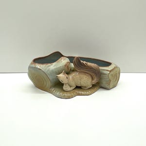 May include: A ceramic planter shaped like a tree log with a squirrel figurine on top. The planter is glazed in shades of brown, green, and blue.