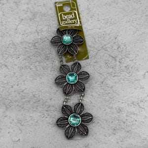 May include: A necklace with three silver-coloured flower pendants, each with a turquoise centre. The flower pendants are linked by a string of small beads. The tag reads "bead gallery".