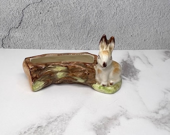 May include: A ceramic planter shaped like a tree stump with a rabbit figurine sitting on the side. The planter is brown and green with a white interior.