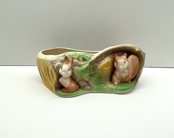 May include: A ceramic planter shaped like a tree trunk with two squirrels peeking out. The planter is painted in shades of brown, green, and yellow.