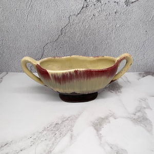 May include: A cream-coloured ceramic serving dish with two handles and a maroon rim. The dish has a scalloped edge and sits on a dark brown base. The exterior has a vertical striped pattern.