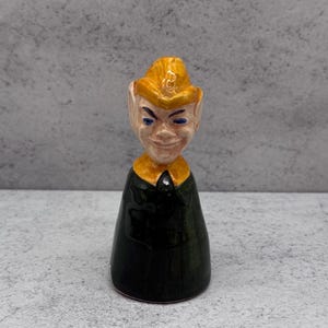May include: A ceramic figurine of a smiling elf with a yellow hat and collar, and a dark green body. The elf's face has blue eyes and a cheerful expression. The figurine is standing on a grey surface.