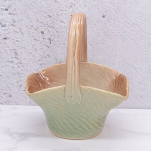May include: A ceramic basket with a light green and brown glaze. The basket has a handle and a textured design.