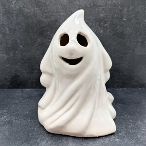May include: White ceramic ghost figurine with a smiling face and two black eyes. The ghost is standing on a dark grey surface.