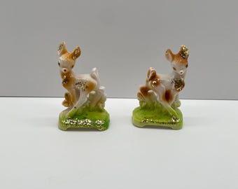 May include: Two small ceramic figurines of deer standing on a green base with gold glitter. The deer are brown and white with gold glitter accents.