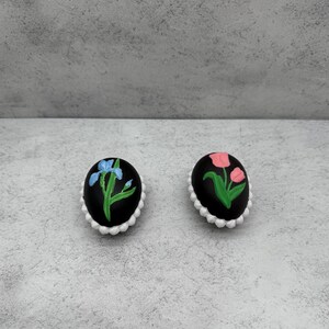 May include: Two black oval decorative items with white scalloped edges. One has a blue iris, the other pink tulips, both with green stems and leaves. These are likely decorative pins or brooches.