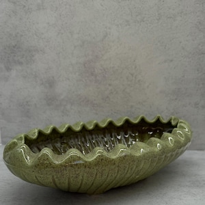 May include: An oval-shaped green ceramic bowl with a wavy edge. The bowl has a speckled texture and a glossy finish. The interior is a darker shade of green. This decorative bowl is suitable for home decor.