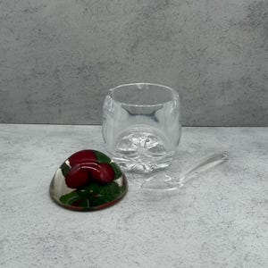May include: A clear glass sugar bowl with a matching spoon and a decorative paperweight featuring red strawberries and green leaves. The sugar bowl has a faceted base.