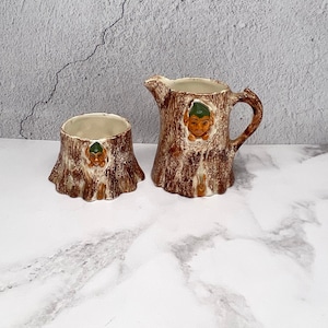 May include: A brown and white ceramic creamer and sugar bowl set shaped like tree stumps with a whimsical face peeking out of each.