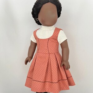 May include: A doll wearing a coral-colored dress with a white turtleneck shirt underneath. The dress has a small floral pattern and a black trim. The doll has black hair and brown skin.