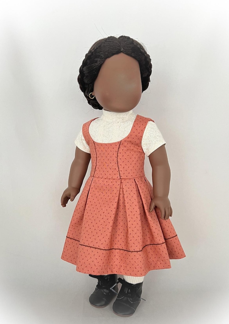 May include: A doll wearing a coral-colored dress with a white turtleneck shirt underneath. The dress has a small floral pattern and a black trim. The doll has black hair and brown skin.