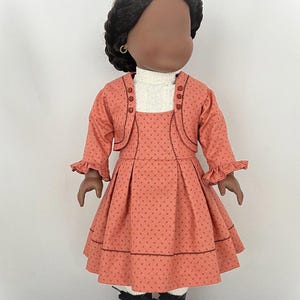 May include: A doll wearing a coral dress with a white turtleneck and a coral jacket. The dress has a small floral pattern and the jacket has buttons down the front. The doll has black hair and brown eyes.