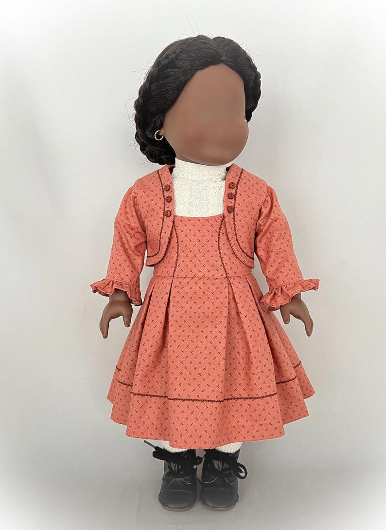 May include: A doll wearing a coral dress with a white turtleneck and a coral jacket. The dress has a small floral pattern and the jacket has buttons down the front. The doll has black hair and brown eyes.