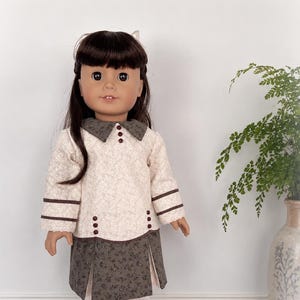 May include: A doll with long dark brown hair wearing a cream-colored long-sleeved top with a small floral pattern, a dark brown collar, and dark brown buttons. The outfit includes a dark brown floral skirt with pleats, white tights, and white boots.