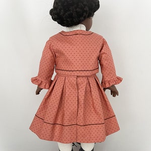 May include: A doll wearing a coral pink dress with a white collar and a matching jacket. The dress has a small floral print and a brown trim. The doll has black hair and is wearing black shoes.
