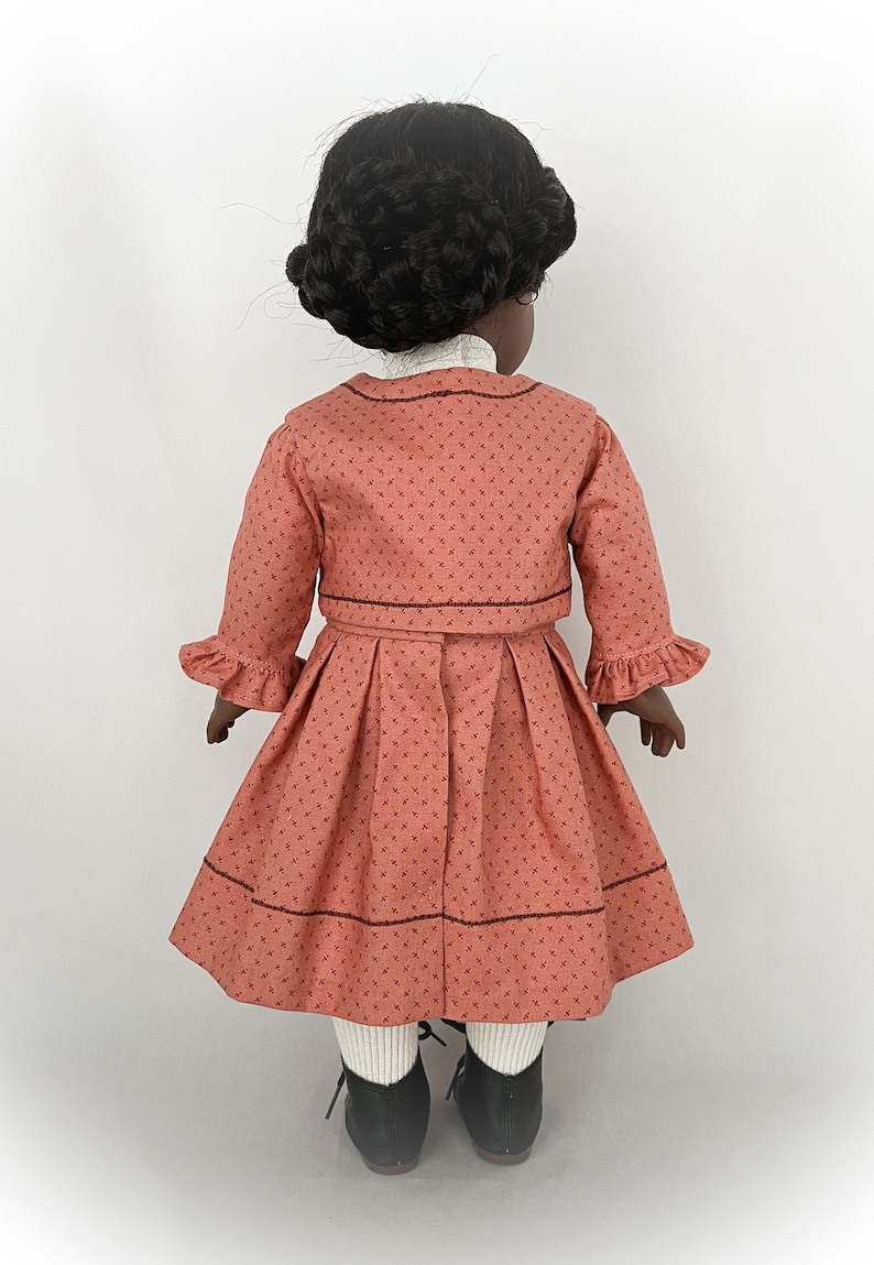 May include: A doll wearing a coral pink dress with a white collar and a matching jacket. The dress has a small floral print and a brown trim. The doll has black hair and is wearing black shoes.
