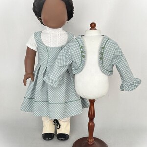 May include: A doll wearing a green and white checkered dress with a green trim and a white blouse underneath. The doll is also wearing white socks and black shoes. A matching green and white checkered jacket is displayed on a wooden stand.