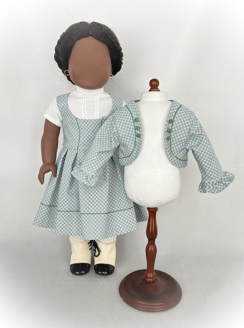 May include: A doll wearing a green and white checkered dress with a green trim and a white blouse underneath. The doll is also wearing white socks and black shoes. A matching green and white checkered jacket is displayed on a wooden stand.