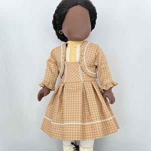 May include: A doll wearing a brown and yellow plaid dress with a white lace trim, a yellow blouse, and a brown jacket with white lace trim. The doll has black hair and brown skin.