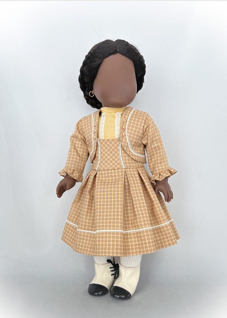 May include: A doll wearing a brown and yellow plaid dress with a white lace trim, a yellow blouse, and a brown jacket with white lace trim. The doll has black hair and brown skin.