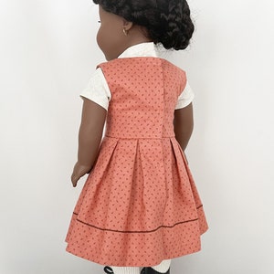 May include: A doll wearing a coral dress with a white undershirt and black shoes. The dress has a small floral pattern and a brown trim around the bottom.