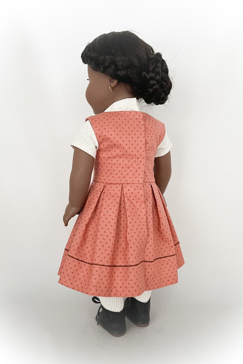 May include: A doll wearing a coral dress with a white undershirt and black shoes. The dress has a small floral pattern and a brown trim around the bottom.