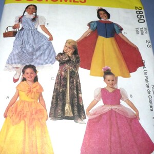 May include: A sewing pattern envelope labeled COSTUMES 2850, size 2-3, featuring illustrations of five different dress designs. The images show a blue gingham dress, a yellow and blue dress with a red cape, a dark patterned gown, a yellow gown, and a pink gown.