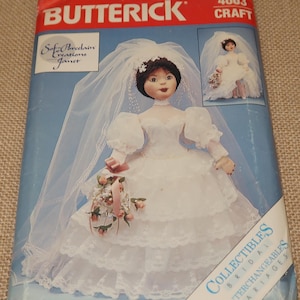 May include: A Butterick craft pattern for a bridal doll. The pattern features a doll in a white wedding dress with a veil and a bouquet. The pattern's packaging is red and blue with the words "Butterick" and "Craft" prominently displayed.