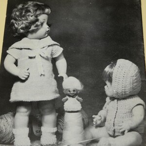 May include: Black and white image of three dolls. The largest doll is standing and wearing a knitted dress and socks. A smaller doll is standing on a spool of thread. The third doll is sitting and wearing a knitted outfit and bonnet.