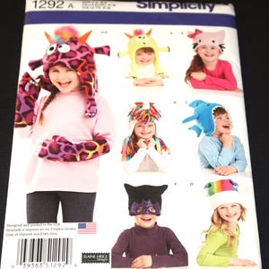 May include: A Simplicity 1292 A sewing pattern envelope featuring illustrations of various animal-themed hats and accessories. The designs include a purple leopard print monster hat with matching mittens, a yellow creature hat, a cat hat, a unicorn hat, a shark hat, a black mask, and a rainbow hat.