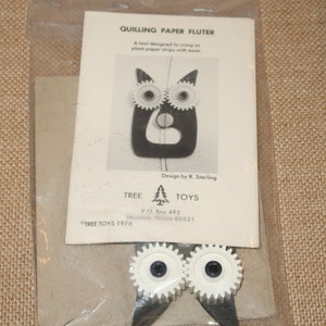 May include: A black and white quilling paper fluter tool designed to crimp or pleat paper strips with ease. The tool is shaped like an owl with two white gears for eyes. The packaging includes the text "Quilling Paper Fluter", "A tool designed to crimp or pleat paper strips with ease", "Design by R. Sterling", "TREE TOYS", "P.O. Box 492", "Hinsdale, Illinois 60521", and "TREE TOYS 1976".