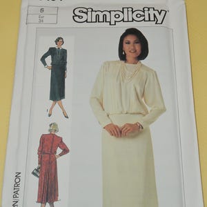 May include: A Simplicity sewing pattern envelope featuring a woman in a cream-colored dress. The envelope includes illustrations of dress designs, the number 7164, and size information. The Simplicity logo is prominently displayed.