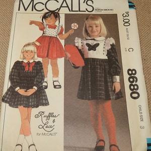 May include: McCall's sewing pattern number 8680 for a child's dress. The pattern includes two different styles, one with a red and white polka dot dress and the other with a plaid dress. The pattern is for child size 3.