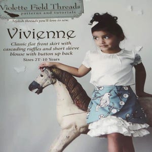 May include: A young girl wearing a white blouse and a blue skirt with a bird and floral print. The skirt has white ruffles. The image includes text that reads "Vivienne" and "Sizes 2T-10 Years". The girl is standing next to a rocking horse.