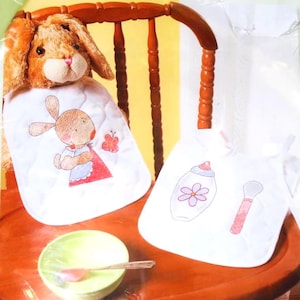 May include: Two white baby bibs with embroidered designs. One bib features a bunny with a red dress and a butterfly. The other bib has a baby bottle and a brush. A green bowl and spoon are also visible.