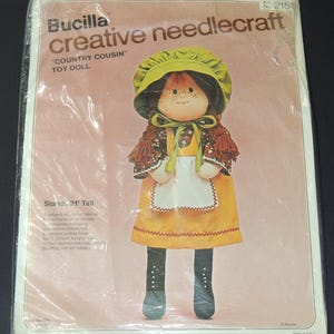 May include: A Bucilla "Country Cousin" toy doll needlecraft kit. The doll stands 21 inches tall and features a yellow dress, green bonnet, and black boots. The kit includes stamped fabric and materials.