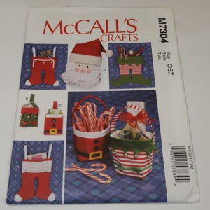 May include: A McCalls Crafts M7304 sewing pattern envelope featuring illustrations of holiday-themed stockings, gift bags, and a Santa face ornament. The designs include red and white fabric elements, green accents, candy canes, and belt details.