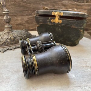 Peut inclure: Lunettes d'opéra vintage noires avec des accents dorés, fermées, reposant sur une surface blanche. Les lunettes sont dans un étui en cuir usé.